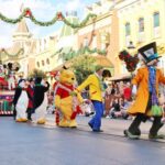 Disney characters, including Winnie the Pooh, Alice, and the Mad Hatter, parade down a festive, decorated street with a large Christmas tree float as crowds watch and take photos.