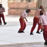 Four performers in red pants and striped shirts act on a white stage outdoors, with a vintage military truck and stone columns in the background. The scene appears to be part of a show or reenactment.