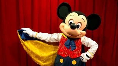 Mickey Mouse in a colorful costume with a red vest, yellow buttons, and a yellow cape, poses in front of a red curtain, smiling and holding out the edge of the cape with one hand.