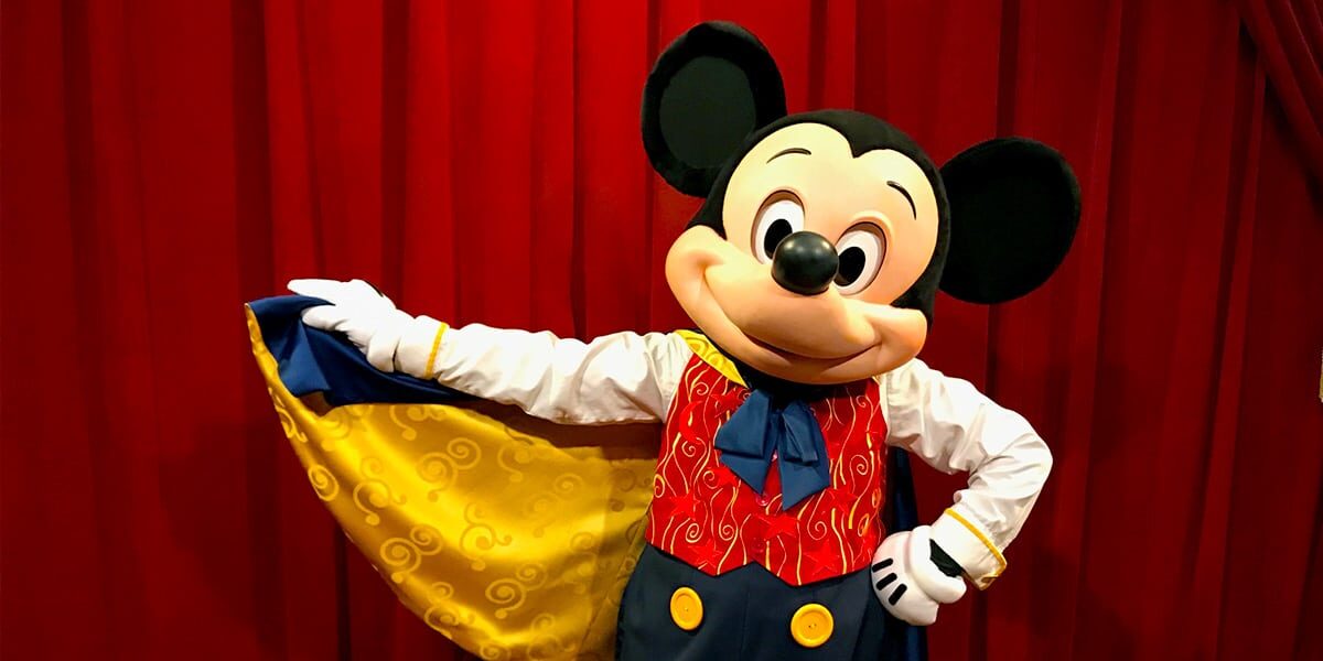 Mickey Mouse in a colorful costume with a red vest, yellow buttons, and a yellow cape, poses in front of a red curtain, smiling and holding out the edge of the cape with one hand.