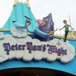 Colorful entrance sign for Peter Pan's Flight features Peter Pan, Wendy, John, and Michael flying over a cloud with the ride’s name in purple letters. A clock tower and blue sky backdrop are also visible.