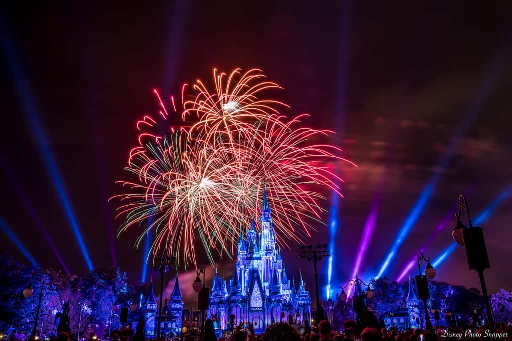 Colorful fireworks burst above a brightly lit Cinderella Castle at night in Disney World, with blue spotlights shining and a crowd watching in the foreground.