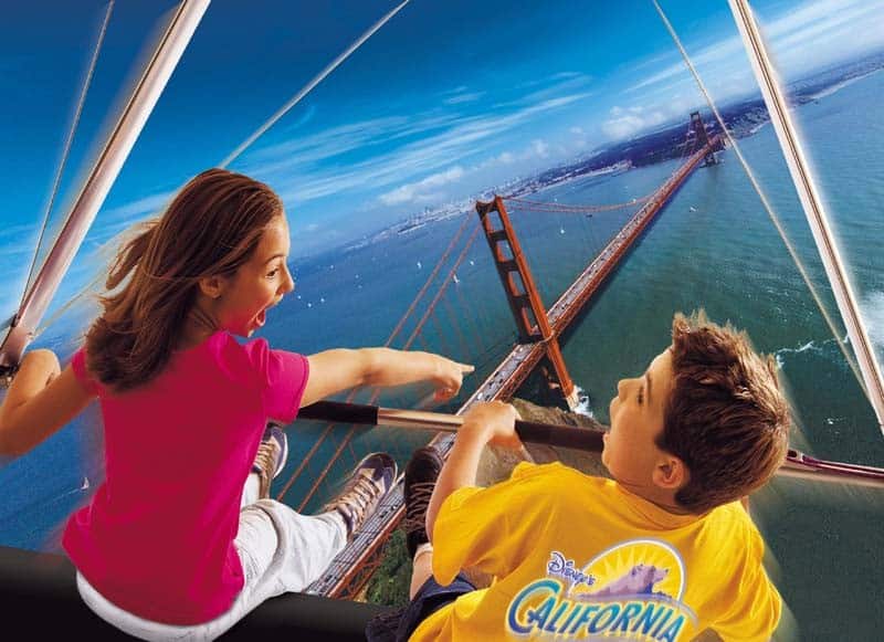 Two children in a virtual hang-glider simulation with a view of the Golden Gate Bridge below. The girl excitedly points at the bridge, while the boy looks on, both appearing thrilled by the experience.