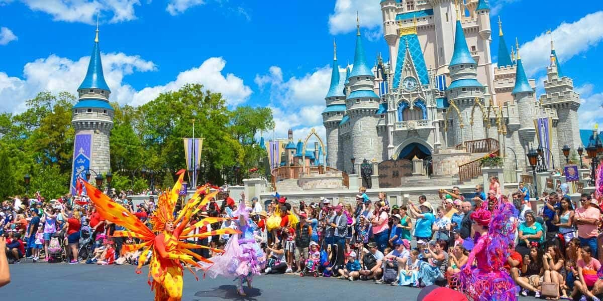 Performers in colorful costumes dance in a parade in front of Cinderella Castle at Disney World, with a large crowd of spectators watching under a bright blue sky.