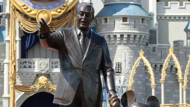 A bronze statue of a smiling man holding hands with Mickey Mouse stands in front of an ornate castle with blue rooftops and golden details at a theme park.