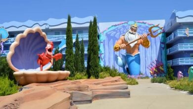 Colorful outdoor area with a large statue of Sebastian the crab in a seashell and a mural of King Triton holding a trident, surrounded by whimsical, ocean-themed hotel buildings and greenery.