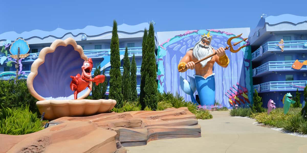 Colorful outdoor area with a large statue of Sebastian the crab in a seashell and a mural of King Triton holding a trident, surrounded by whimsical, ocean-themed hotel buildings and greenery.