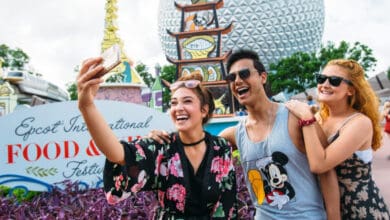 Three young adults smile and take a selfie together at the Epcot International Food & Wine Festival, one of the popular Disney World events, with colorful signs, garden plants, and the iconic Epcot sphere in the background.