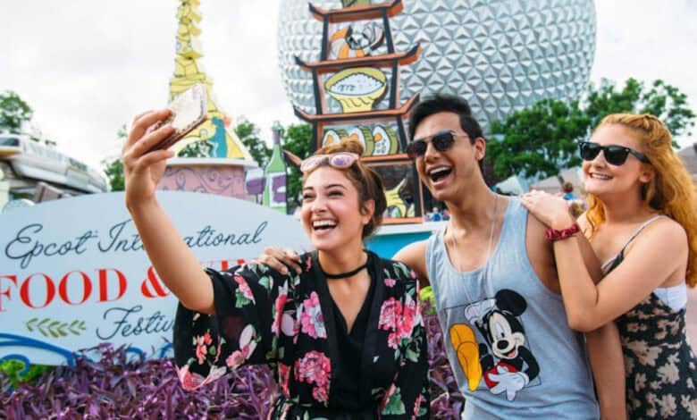 Three young adults smile and take a selfie together at the Epcot International Food & Wine Festival, one of the popular Disney World events, with colorful signs, garden plants, and the iconic Epcot sphere in the background.