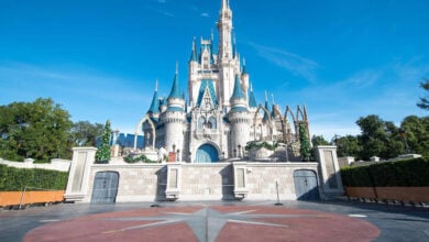 Cinderella Castle at Walt Disney World, decorated with garlands, stands under a clear blue sky. The foreground shows an empty plaza with a star pattern on the ground.