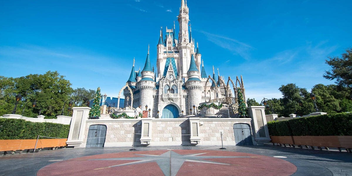 Cinderella Castle at Walt Disney World, decorated with garlands, stands under a clear blue sky. The foreground shows an empty plaza with a star pattern on the ground.