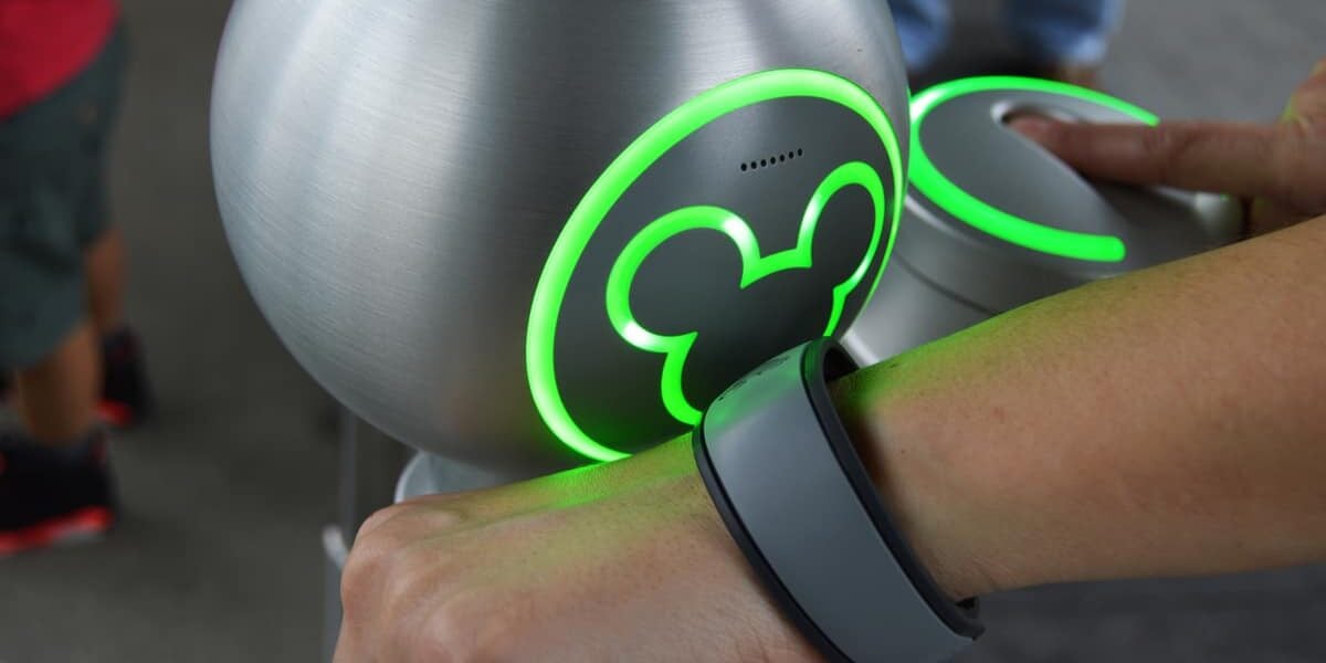A person scans a gray MagicBand on their wrist at a glowing green Mickey Mouse reader at a Disney park entrance.