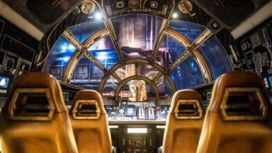 A futuristic spacecraft cockpit with two brown pilot seats facing a control panel filled with buttons and screens. Large transparent windows provide a view of a space-themed setting outside.