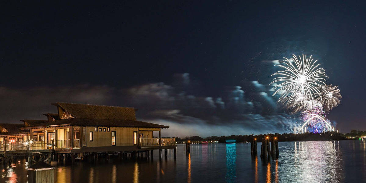 A nighttime lakeside scene with an overwater bungalow on the left and bright fireworks lighting up the sky above a distant theme park across the water. Reflections shimmer on the calm surface of the lake.