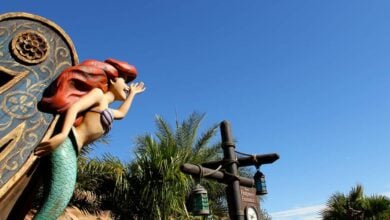 A statue of Ariel, the Little Mermaid, with red hair and a green tail, appears to be singing atop a decorative structure, set against a bright blue sky and palm trees.