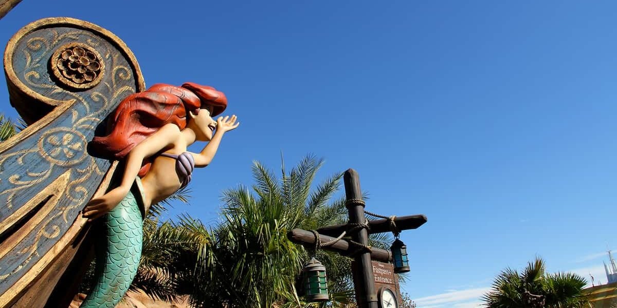 A statue of Ariel, the Little Mermaid, with red hair and a green tail, appears to be singing atop a decorative structure, set against a bright blue sky and palm trees.