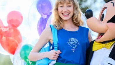 A smiling woman with blonde curly hair wearing a blue tank top and backpack stands next to a person in a Mickey Mouse costume, with colorful Mickey-shaped balloons in the background—perfect for capturing those Magic Kingdom Tips moments.