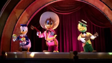 Three colorful animatronic characters—Donald Duck with maracas, José Carioca with a guitar, and Panchito Pistoles in a sombrero—stand on stage under a decorative arch with red curtains, delighting fans amid recent Walt Disney World rumors.