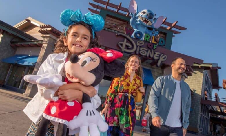 A smiling girl with blue Minnie Mouse ears holds a Minnie Mouse plush in front of the World of Disney store at Disney Springs, as two adults happily walk behind her under a clear blue sky at Disney World.