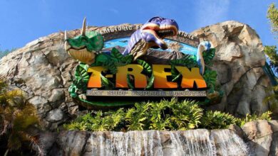 Sign for the T-REX restaurant features large dinosaur models, including a green triceratops and a purple T-Rex, atop rocky terrain with greenery and a cascading waterfall beneath a blue sky.