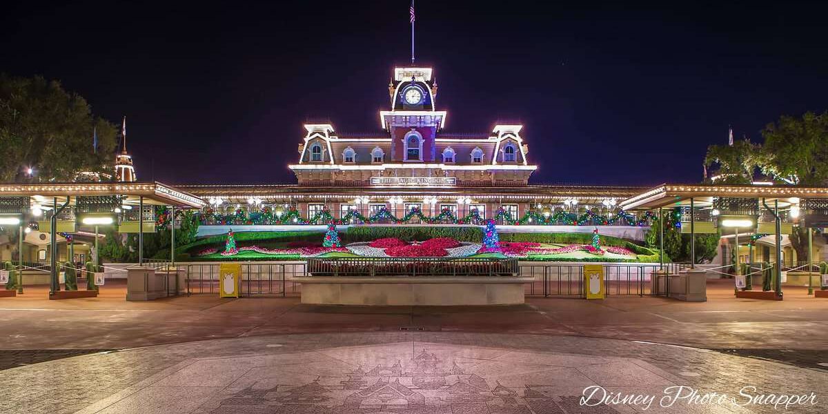 The entrance to Magic Kingdom at Walt Disney World is brightly lit at night, featuring the Main Street Train Station and colorful Mickey Mouse-shaped flowerbeds in front.