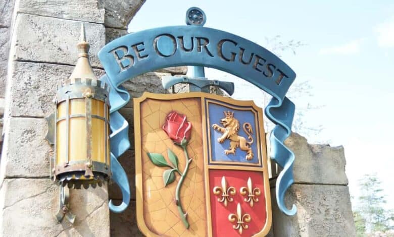 A decorative sign reading "Be Our Guest" is displayed on a stone wall. Below the text, a shield features a rose on the left and a lion with fleur-de-lis on the right. A vintage lantern hangs nearby. The sky is partly visible at Disney World.