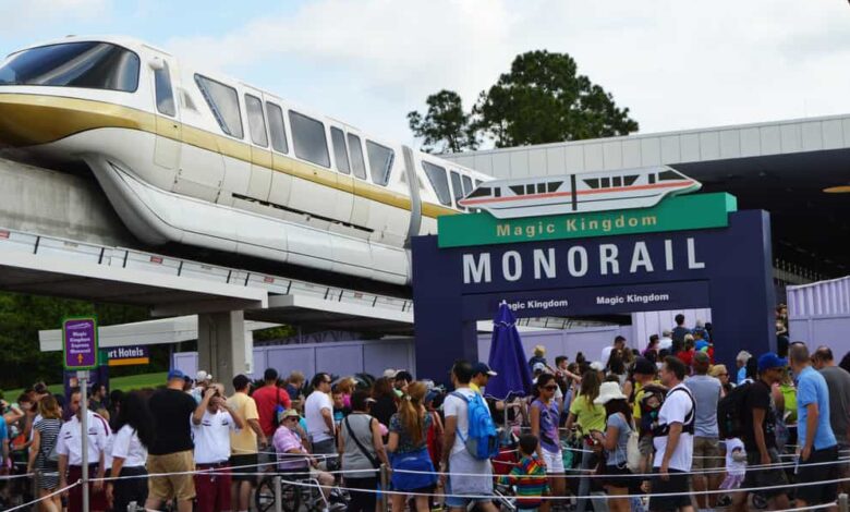 A monorail glides above a crowded entrance to the Magic Kingdom Monorail station, with many people waiting in line under a sign that reads "Magic Kingdom Monorail" on a sunny day.