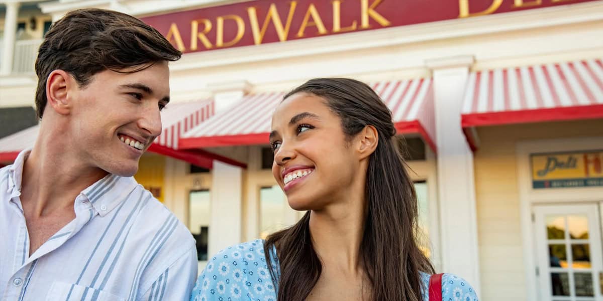 A young man and woman smile at each other while standing outside Disney’s BoardWalk Villas, with red-and-white striped awnings and a "BoardWalk Deli" sign in the background.