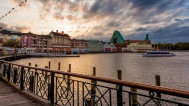 A scenic boardwalk with decorative railing curves along a waterfront at Disney’s BoardWalk Villas, with colorful buildings and a unique teal-roofed hotel in the background under a dramatic, cloudy sunset sky. A boat floats on the calm water.