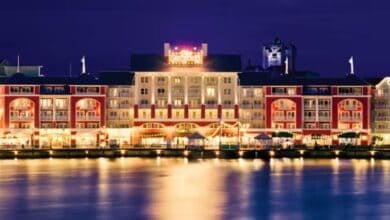 A brightly lit hotel with a mix of red and beige exteriors reflects on a calm body of water at night. The warm lights create a welcoming atmosphere, with a tower visible in the background against the night sky.