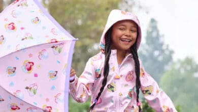 A young girl wearing a pink raincoat with cartoon princess prints smiles while holding a matching pink Disney Umbrella