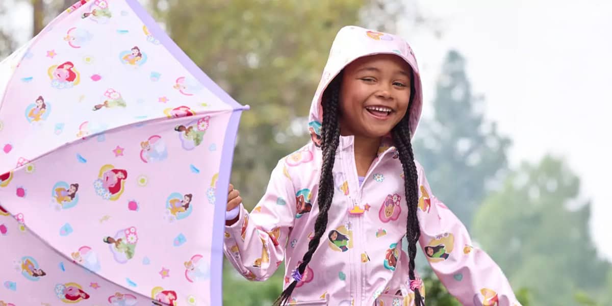 A young girl wearing a pink raincoat with cartoon princess prints smiles while holding a matching pink Disney Umbrella