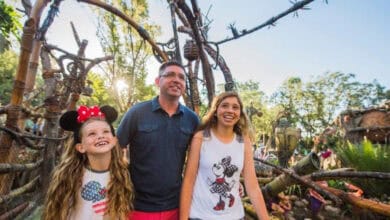 A smiling man and two girls, one wearing Minnie Mouse ears, stand at Pandora – The World of Avatar, enjoying a bright, sunny day.