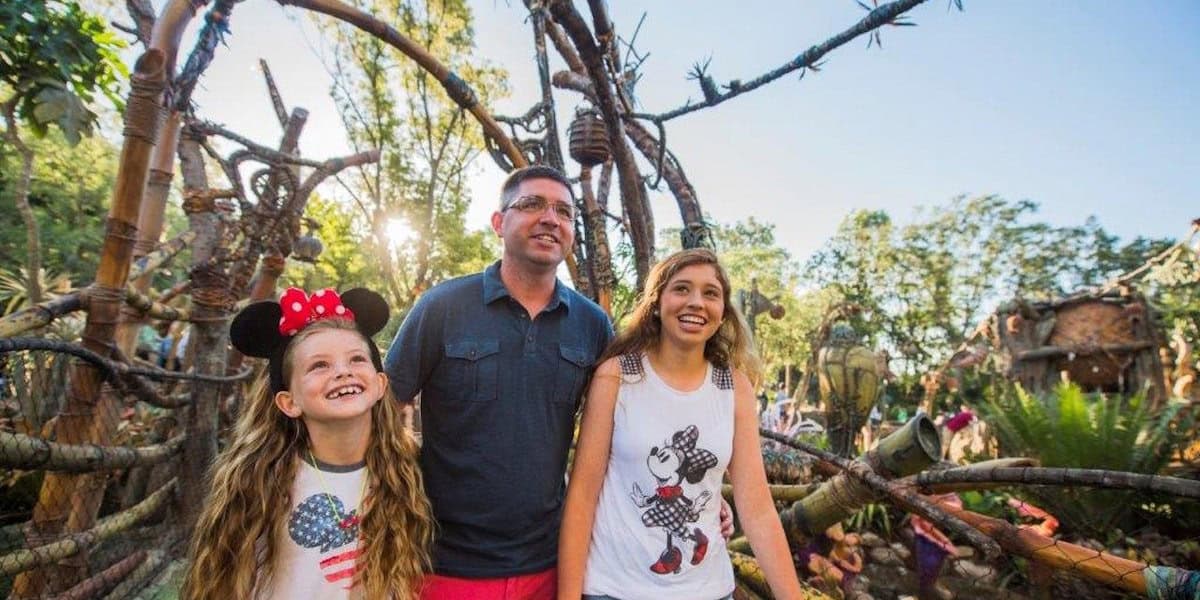 A smiling man and two girls, one wearing Minnie Mouse ears, stand at Pandora – The World of Avatar, enjoying a bright, sunny day.