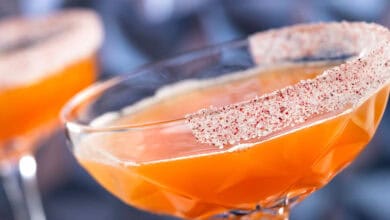 A close-up of a vibrant non-alcoholic drink in an orange hue, served in a glass with a patterned rim coated in red and white spiced powder, with another similar beverage blurred in the background at Disneyland Park.