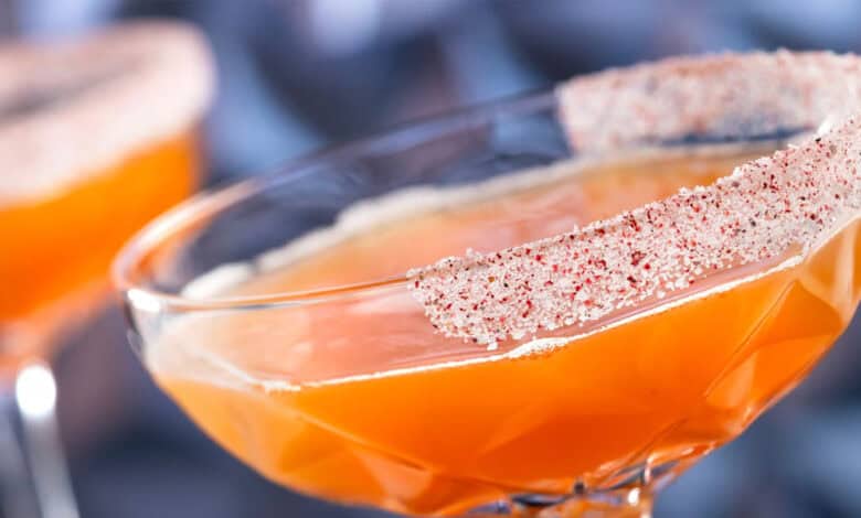 A close-up of a vibrant non-alcoholic drink in an orange hue, served in a glass with a patterned rim coated in red and white spiced powder, with another similar beverage blurred in the background at Disneyland Park.