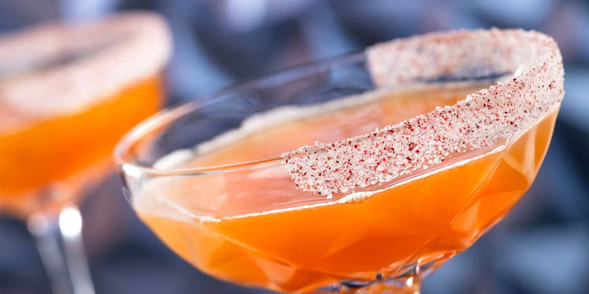 A close-up of a vibrant non-alcoholic drink in an orange hue, served in a glass with a patterned rim coated in red and white spiced powder, with another similar beverage blurred in the background at Disneyland Park.