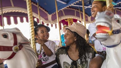 A woman and two children smile and laugh while riding white carousel horses under a brightly colored canopy, enjoying a fun Mother-Son Day at Walt Disney World together.