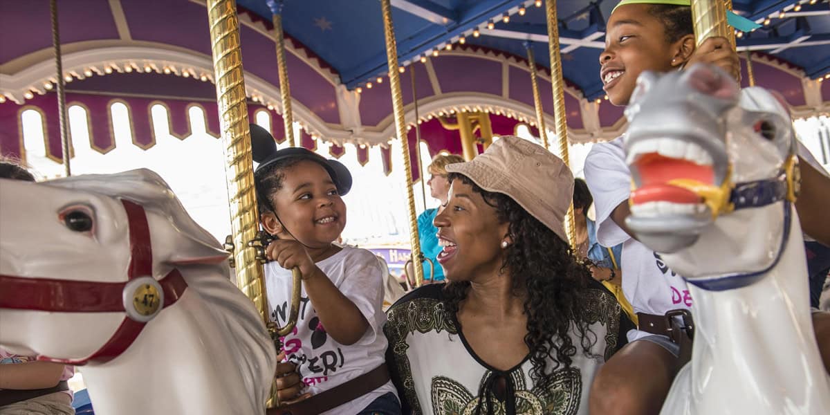 A woman and two children smile and laugh while riding white carousel horses under a brightly colored canopy, enjoying a fun Mother-Son Day at Walt Disney World together.