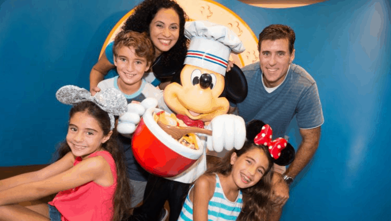 A smiling family with two adults and three children, wearing Disney-themed mouse ears, poses with a person in a chef Mickey Mouse costume against a colorful background.