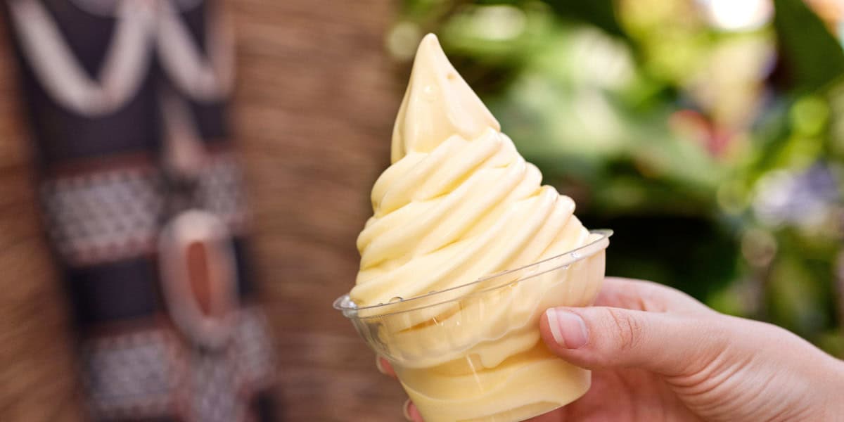 A hand holds a cup of creamy, yellow soft-serve ice cream with a swirled top; the background features greenery and a tiki-style design, capturing the fun of vegan meals at Disneyland.