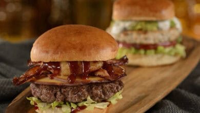 Two gourmet burgers on a wooden platter; the front burger boasts lettuce, a beef patty, cheese, bacon, onion rings, and barbecue sauce—just what you’d expect from the best burgers at Disney World. The back features lettuce, tomato, and a lighter patty.