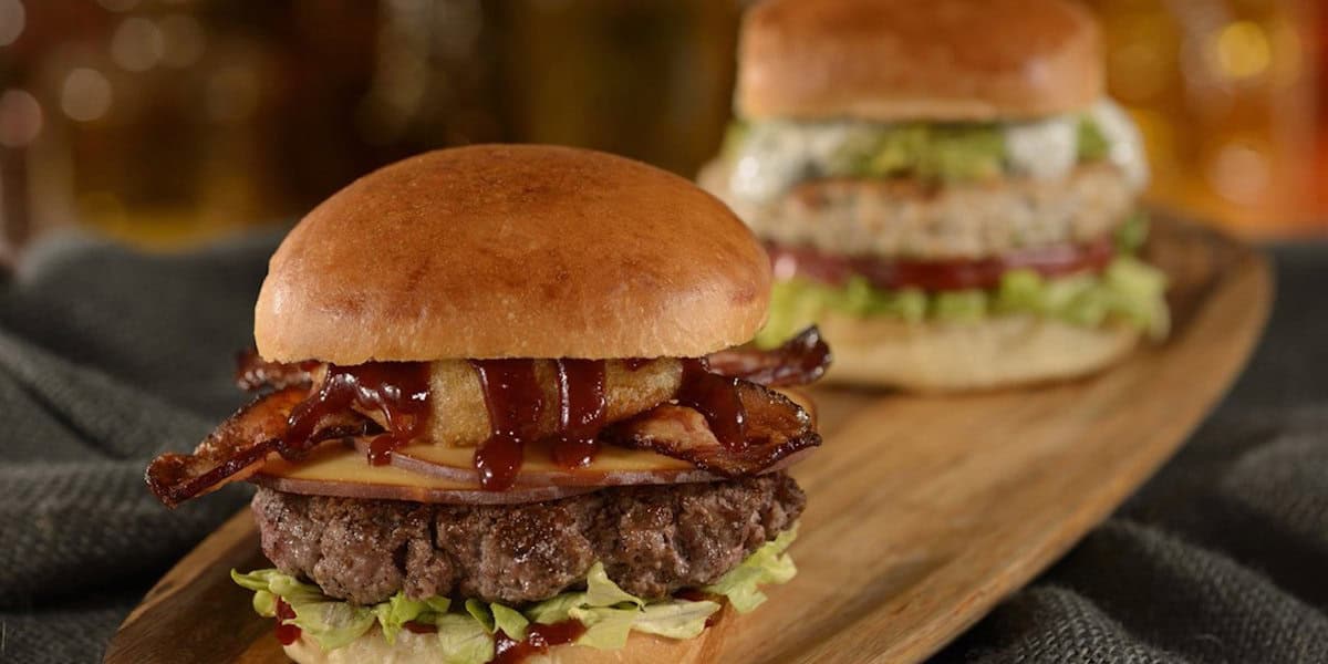 Two gourmet burgers on a wooden platter; the front burger boasts lettuce, a beef patty, cheese, bacon, onion rings, and barbecue sauce—just what you’d expect from the best burgers at Disney World. The back features lettuce, tomato, and a lighter patty.