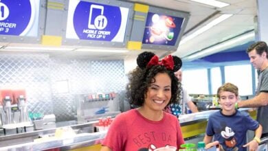 A woman wearing Minnie Mouse ears and a red shirt smiles while holding a tray of food in a casual restaurant, enjoying the ease of Mobile Ordering. A young boy and another adult are behind her under digital signs reading “Mobile Order Pick Up.”.