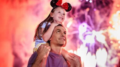 A man carries his smiling daughter on his shoulders during a Daddy Daughter Disney Day. The girl, wearing Minnie Mouse ears, looks excited as colorful fireworks light up the sky behind them.