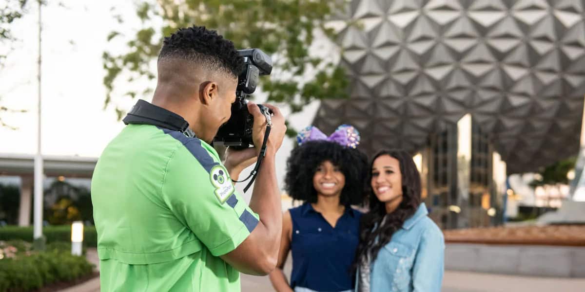 A photographer in a green shirt snaps a photo of two smiling women, one in mouse ears, in front of the iconic geodesic sphere at Epcot—a perfect way to capture one of the EPCOT must-do experiences at this Disney theme park.