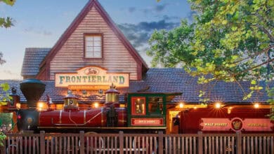 A vintage red steam locomotive is parked in front of a wooden Frontierland Railroad Station building, surrounded by green trees and illuminated with warm lights.