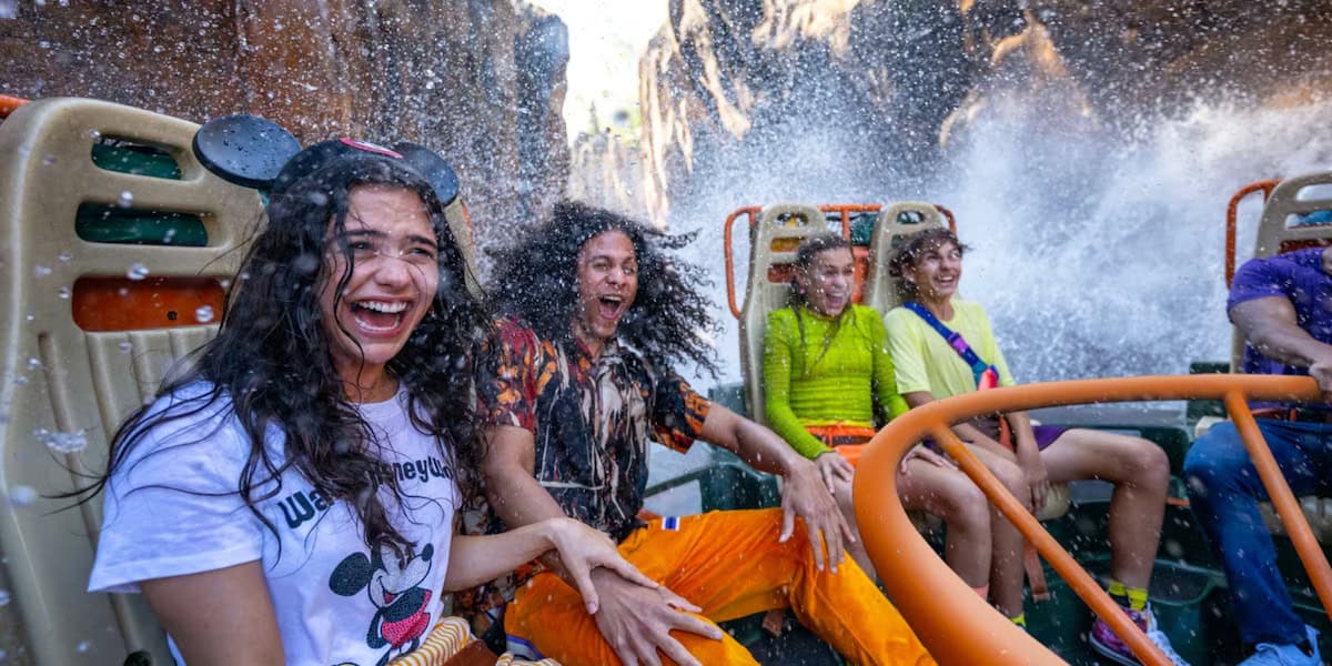 Four people laugh and get soaked by splashing water while riding a theme park water ride in Asia at Disney’s Animal Kingdom, surrounded by rocky scenery. One woman sports Mickey Mouse ears and a Mickey shirt.