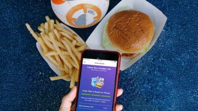 A hand holds a smartphone displaying a Mobile Ordering app. Next to it are a tray of French fries, a burger in a paper container, and a BB-8 droid cup on a blue speckled surface.