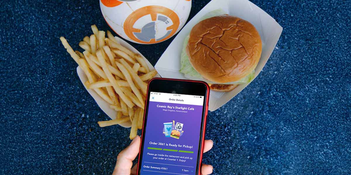 A hand holds a smartphone displaying a Mobile Ordering app. Next to it are a tray of French fries, a burger in a paper container, and a BB-8 droid cup on a blue speckled surface.